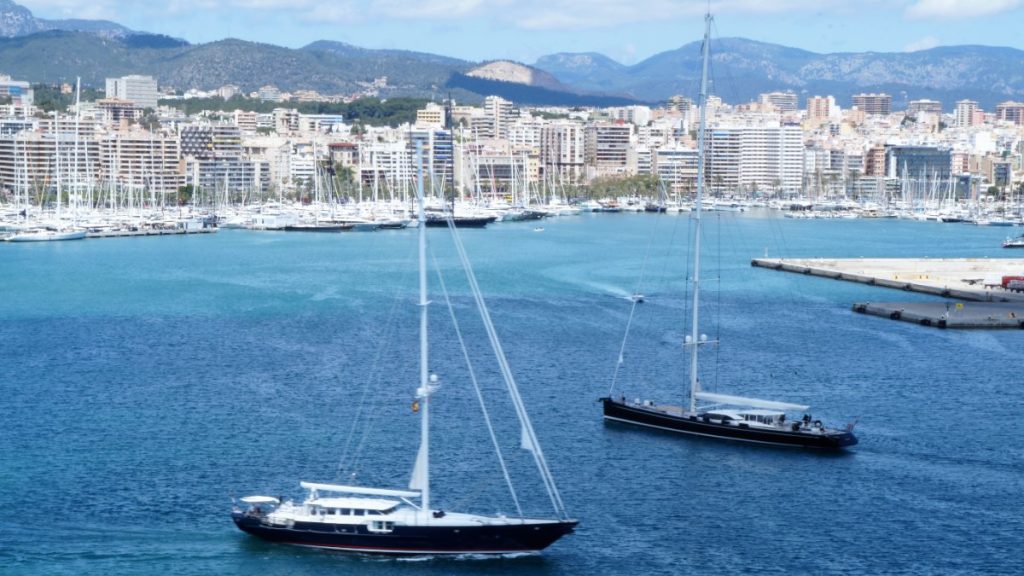 Dos grandes veleros navegan por la bahía de Palma con el puerto deportivo y la ciudad al fondo