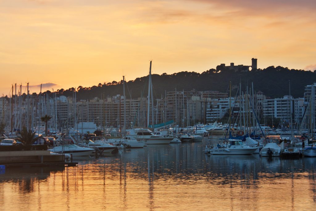 Embarcaciones amarradas en el puerto de Palma al atardecer, con el castillo de Bellver al fondo