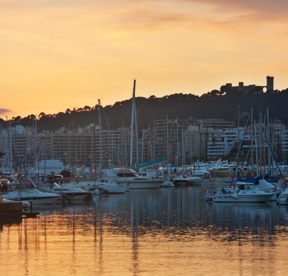 Embarcaciones amarradas en el puerto de Palma al atardecer, con el castillo de Bellver al fondo
