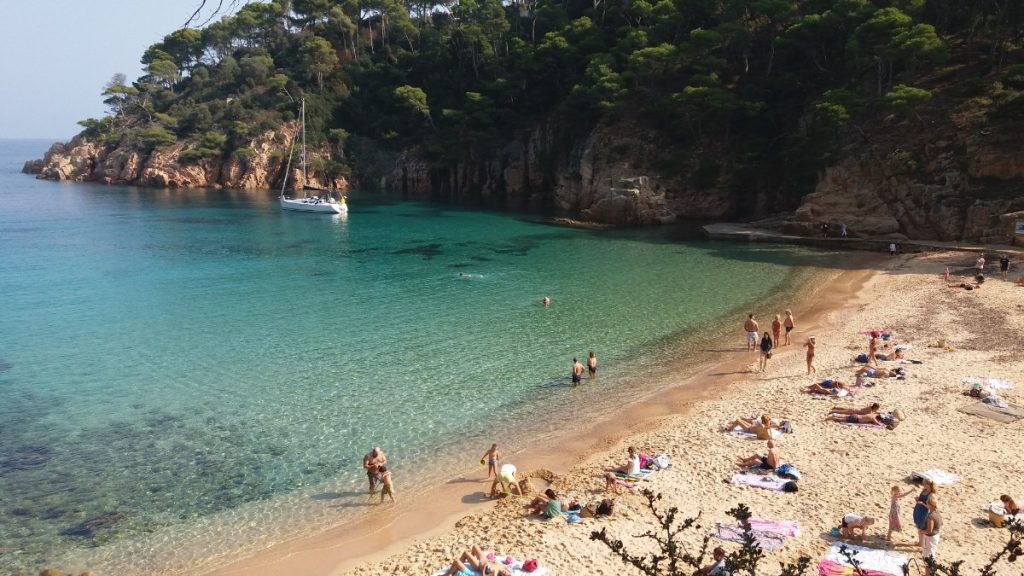 Playa de la cala de Aiguablava, en Begur, con bañistas, aguas turquesa, pinos sobre las rocas y un velero fondeado al fondo