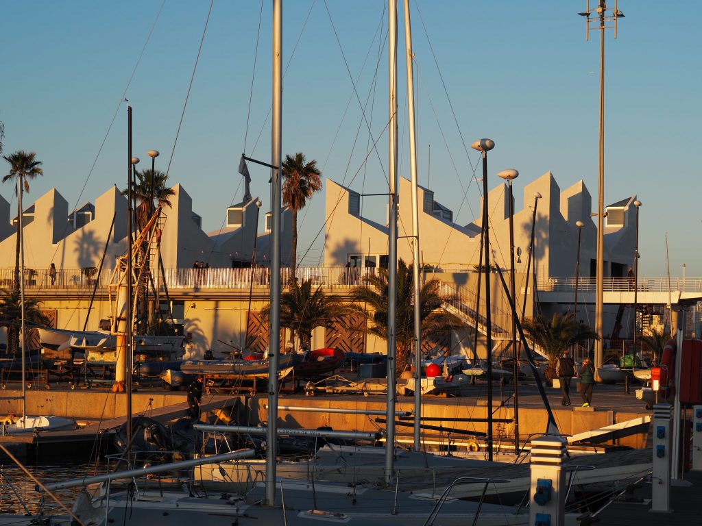 Edificio del Centre Municipal de Vela de Barcelona en el Port Olímpic, con embarcaciones y mástiles en primer plano al atardecer