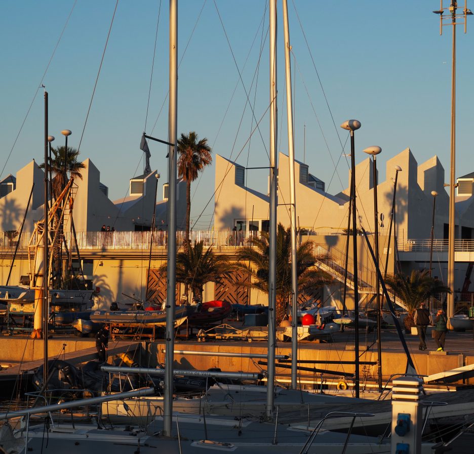 Edificio del Centre Municipal de Vela de Barcelona en el Port Olímpic, con embarcaciones y mástiles en primer plano al atardecer