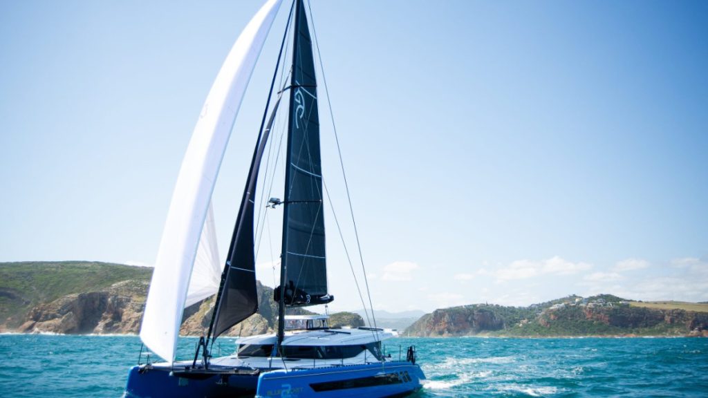 El catamarán Knysna 550, de casco azul, navegando con mayor negra y vela de proa blanca junto a una costa de acantilados