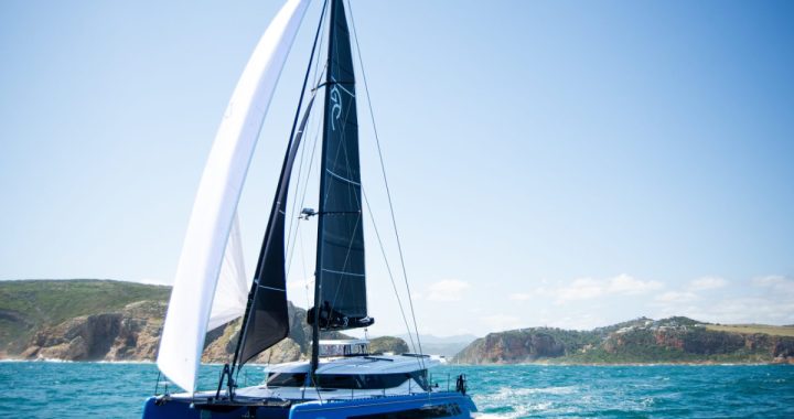 El catamarán Knysna 550, de casco azul, navegando con mayor negra y vela de proa blanca junto a una costa de acantilados