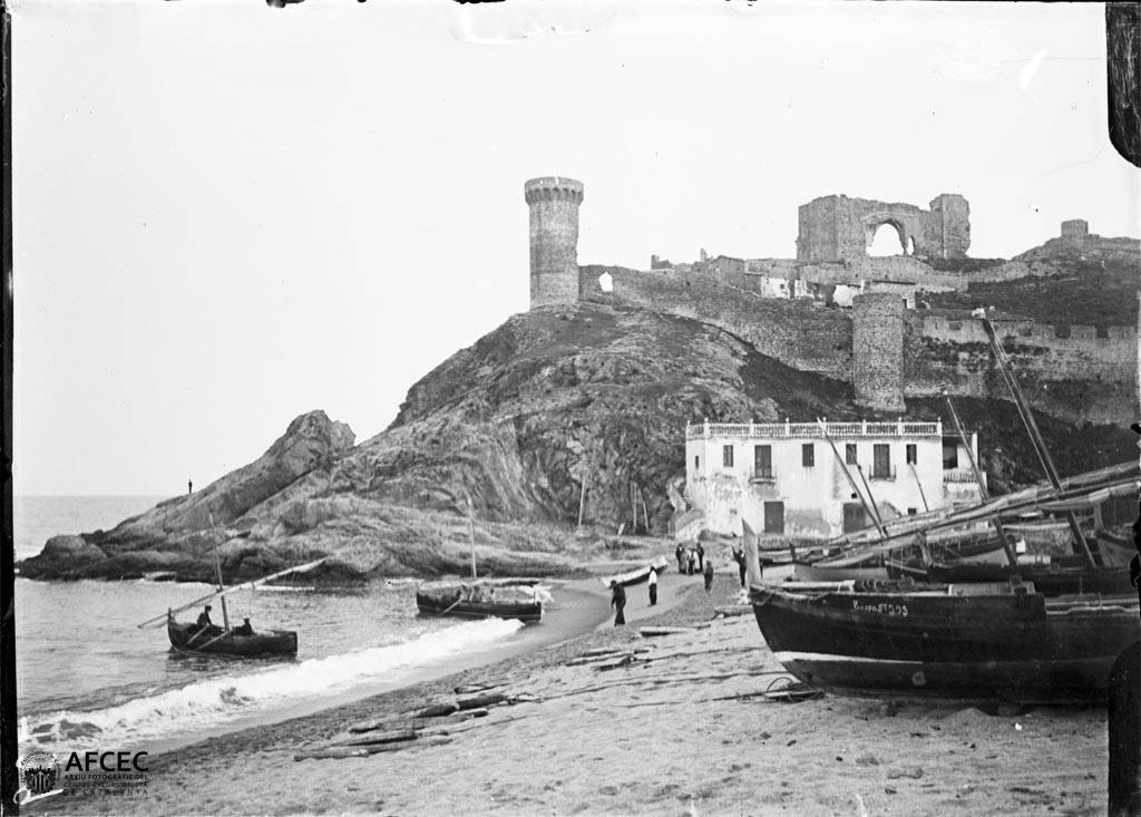 Fotografía antigua de barcas en la playa de Tossa de Mar, algunas con su antena de vela latina, con la torre y las murallas de la Vila Vella al fondo